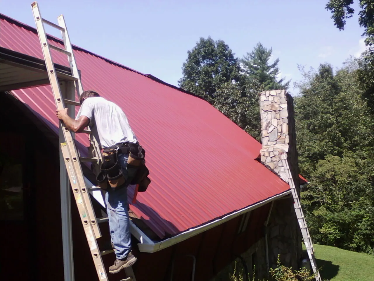 Roofer on a ladder beside a red metal roof and stone chimney — Soffit & Fascia Repair by Roofing Brothers in Lagro